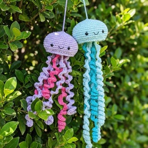 May include: Two handmade crochet jellyfish ornaments. One is purple with pink and lavender tentacles, the other is light blue with blue tentacles. Both have smiling faces and are hanging from a green bush.