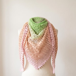 May include: A handmade crochet shawl in a triangle shape, displayed on a mannequin. The shawl features a gradient of colors, starting with green at the top, transitioning to peach, and ending with pink. The open weave design is visible.
