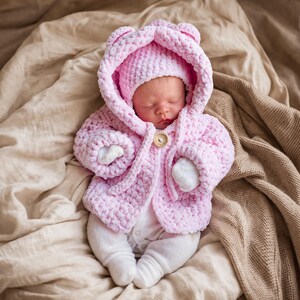 Puede incluir: Un bebé dormido con una chaqueta con capucha rosa de ganchillo y un gorro a juego. La chaqueta tiene orejas de oso y un cierre de botón de madera. El bebé también lleva un body y calcetines blancos, tumbado sobre una manta beige.