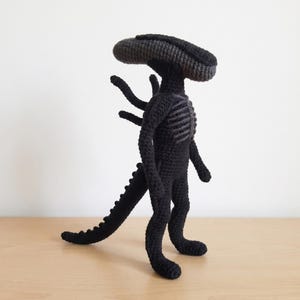 Op de afbeelding: Een handgemaakte, zwarte gehaakte Alien-figuur met een lange, gesegmenteerde staart en een geribbelde borst. Het wezen heeft een grote, langwerpige kop en vier gebogen aanhangsels. De figuur staat op een houten oppervlak.