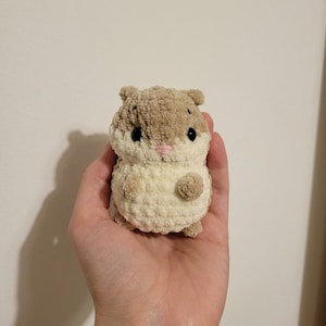 May include: A handmade crochet hamster toy in shades of cream and brown. The plush toy has black eyes, a pink nose, and small, rounded ears. The hamster is held in a hand, showcasing its soft, textured appearance.