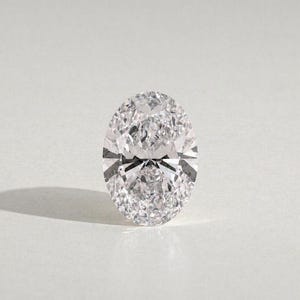 Può includere: Una gemma trasparente e incolore con taglio ovale e superficie sfaccettata brillante. La pietra presenta una disposizione simmetrica di sfaccettature triangolari e a forma di aquilone che riflettono la luce creando un effetto scintillante.