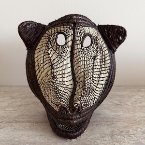 May include: A woven mask shaped like a primate head, featuring dark brown and cream-colored fibers. The mask has two circular eye openings, prominent ears, and a defined snout area, all constructed with a detailed, textured coil weave pattern.