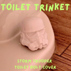 Peut inclure: Un cache-boulon de toilette en plastique blanc en forme de casque de Stormtrooper. Le texte TOILET TRINKET figure en haut et STORM TROOPER TOILET BOLT COVER en bas. Le cache est placé sur un boulon sur une base de toilette blanche.