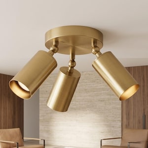 Op de afbeelding: Een plafondlamp met drie lichtpunten, een ronde basis en drie verstelbare cilindrische spots in een geborstelde messing afwerking. Elke lichtkop is verbonden met de centrale houder via een draaibaar gewricht voor gerichte verlichting.
