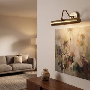 May include: A brass picture light illuminates an abstract painting on a wall. The painting features a blend of colors, including green, purple, and beige. A small vase sits on a wooden cabinet below the artwork. A sofa and rug are visible in the background.