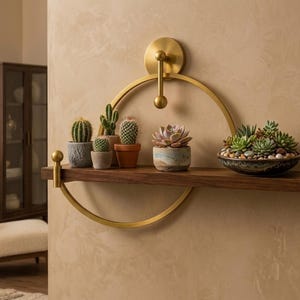 May include: A wall-mounted shelf featuring a dark brown wooden plank supported by a circular gold metal frame. The shelf holds several small potted cacti, succulents, and a shallow bowl filled with various small succulent plants and pebbles.