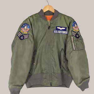 May include: Olive green bomber jacket with gray ribbed cuffs and collar. Features patches on the sleeves and chest, including "TOMCAT" and "U.S. AIR FORCE" text. The jacket has a zipper closure and multiple pockets.