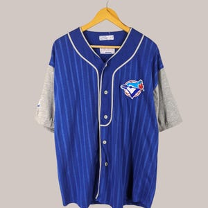 May include: Blue baseball jersey with white pinstripes and grey sleeves. Features a team logo on the chest and a button-up front. The jersey is hanging on a wooden hanger.