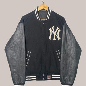 Può includere: Giacca college nera con maniche in pelle. Presenta il logo dei New York Yankees sul petto. La giacca ha strisce bianche sul colletto, sui polsini e sull'orlo. Le maniche sono di colore grigio scuro.