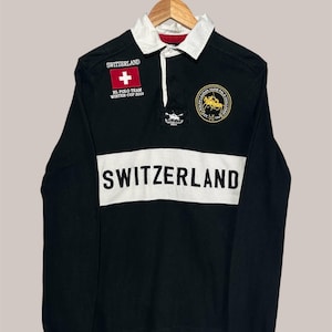 Puede incluir: Camiseta de rugby negra de manga larga con cuello blanco y una franja horizontal blanca en el pecho. La palabra "SWITZERLAND" está impresa en blanco. Incluye un parche de la bandera suiza y un emblema dorado.
