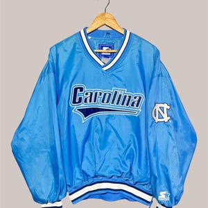 May include: Light blue Carolina pullover jacket with white and navy accents. Features the word "Carolina" in large lettering across the chest, along with a "NC" logo on the sleeve. The jacket has a V-neck and elastic cuffs.