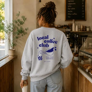Könnte beinhalten: Ein hellgraues Sweatshirt mit Rundhalsausschnitt und einem dunkelblauen Aufdruck auf der Rückseite. Das Design zeigt den Text "local coffee club", eine Hand, die eine Kaffeetasse hält, sowie die Sätze "FUEL YOUR HAPPINESS" und "take a break".