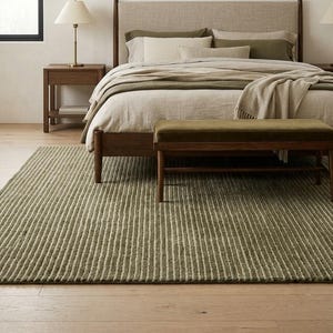 May include: A bedroom featuring a wooden bed frame with a beige upholstered headboard, layered with olive green and cream bedding. A matching wooden bench sits at the foot of the bed on a textured olive green striped rug, flanked by two nightstands.
