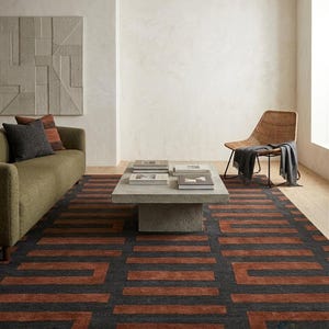 May include: A rectangular area rug featuring a repeating geometric pattern of dark charcoal gray and burnt orange horizontal bars. The rug is positioned on a light wood floor, extending underneath a concrete coffee table and an olive green sofa.