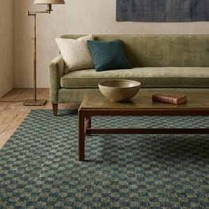 May include: A rectangular area rug featuring a repeating checkerboard pattern in alternating shades of dark green and muted blue. The rug is positioned on a wooden floor beneath a low wooden coffee table, a velvet sofa, and a floor lamp.