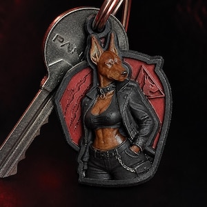 Puede incluir: Llavero detallado con un Doberman Pinscher con físico humanoide, vistiendo una chaqueta de cuero negra y jeans. El llavero tiene un diseño de escudo rojo y negro y está unido a una llave plateada.
