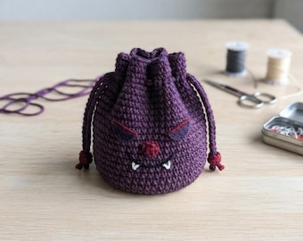 May include: A purple crochet drawstring pouch featuring a monster face design with dark purple felt eyes, a red nose, and white embroidered fangs. The bag has a round base and two matching purple braided drawstrings with knotted ends for closure.