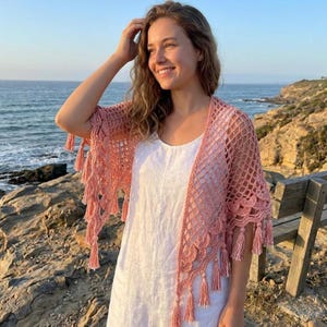 Peut inclure: Un châle en crochet rose poudré avec un motif en losanges ajouré, des bords festonnés et des pompons suspendus le long de l'ourlet. Le vêtement est porté sur une robe blanche sans manches, mettant en valeur le travail du fil.