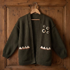 May include: An olive green crochet cardigan featuring long sleeves, a ribbed collar, and ribbed cuffs. The front displays four circular eye appliqués on the left chest and two pockets at the bottom, each accented with white triangular teeth.