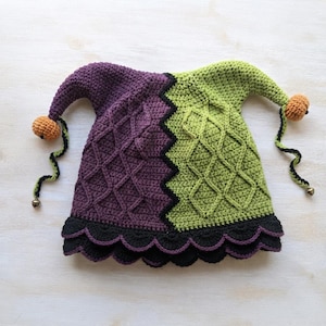 May include: A two-toned jester hat crocheted with purple and lime green yarn featuring a diamond cable stitch pattern. The hat has two pointed ends tipped with orange pom-poms and small bells, finished with a black scalloped edge at the base.