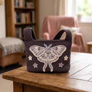 May include: A dark purple crochet bag featuring a large, detailed moth appliqué in shades of cream, light purple, and grey. The moth design includes a crescent moon on its body. Small, crocheted stars in cream and light purple surround the moth.