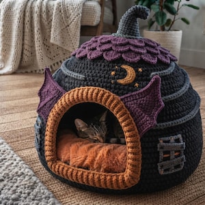 May include: A dark grey crocheted pet cave shaped like a witch's hat with purple scale-like details, a yellow crescent moon embroidery, and purple bat-like wings flanking the orange-trimmed entrance, containing a sleeping tabby cat on a plush orange bed.