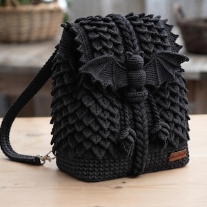 May include: A black crochet backpack featuring a textured scale-like pattern across the exterior. The front flap is adorned with a three-dimensional crocheted bat figure with spread wings. A small brown rectangular tag is attached to the side.