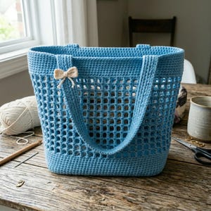 May include: A light blue crochet tote bag with a rectangular open weave design. The bag has two handles and a small beige bow attached to one of the handles. The bag is sitting on a wooden table.