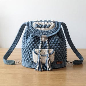 Peut inclure: Un sac à dos en crochet bleu et crème avec un motif texturé en points noisette, un rabat orné d'une tresse, une poche avant à bouton-pression, deux cordons tressés avec pompons et des bretelles réglables munies de clips métalliques.