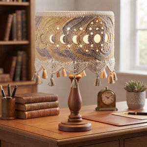 May include: A table lamp with a wooden base and a crocheted lampshade featuring a moon phase pattern and star-shaped cutouts. The shade is decorated with small hanging tassels along the bottom edge and emits a warm light through the fabric.