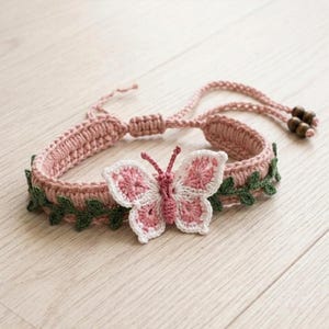 May include: A crochet bracelet featuring a dusty pink band adorned with small green leaf-like accents and a central butterfly motif in shades of pink and white. The adjustable band includes two dark brown wooden beads at the ends of the tie strings.