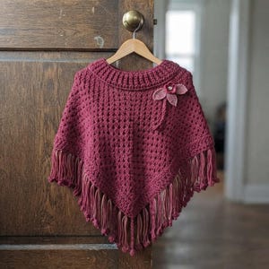May include: A deep maroon crochet poncho featuring a textured stitch pattern, a ribbed neckline, and long fringe along the bottom hem. A small, decorative crochet flower with three petals is attached to the upper right side of the garment.