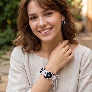 May include: A white crochet bracelet featuring black floral motifs with central white beads. The bracelet is constructed from white thread and includes two dangling white strings, each finished with a small white bead at the end for closure.