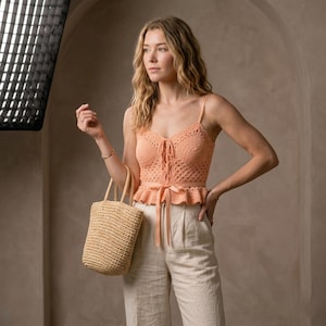 May include: A peach-colored crochet top with spaghetti straps and a tie-up front. The top has a ruffled hem and is paired with beige linen pants. A woven straw tote bag is held in the hand.