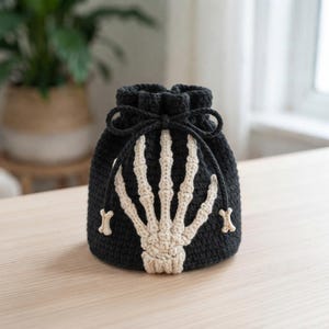 Op de afbeelding: Een zwart gehaakt buideltasje met trekkoord en een crèmekleurig skelet-handontwerp op de voorzijde. De tas heeft een bijpassend zwart trekkoord met kleine botvormige kralen aan de uiteinden, wat zorgt voor een gedetailleerd uiterlijk.