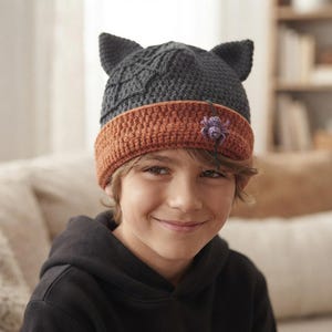 May include: A dark gray crocheted beanie with two pointed cat ears on top. The hat features a textured spiderweb pattern on the side and a wide, folded orange brim. A small, purple crocheted spider is attached to the front of the orange brim.