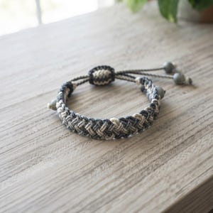 May include: A braided cord bracelet featuring a mix of charcoal gray, light gray, and cream-colored threads. The design includes a sliding knot closure and two dangling cord ends, each finished with a small, smooth, round gray bead.
