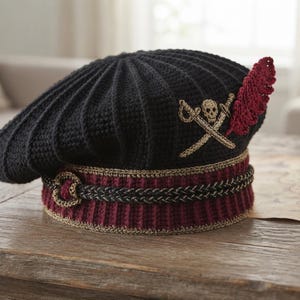 May include: A black crocheted beret featuring a maroon ribbed brim with gold trim. The hat is decorated with an embroidered gold skull and crossbones emblem and a single red crocheted feather accent attached to the side of the crown.