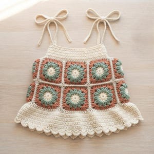 May include: A sleeveless crochet top featuring a square-patterned bodice with cream, sage green, and terracotta floral motifs. The garment has tie-up shoulder straps and a flared, scalloped hemline, all crafted from cream-colored yarn.