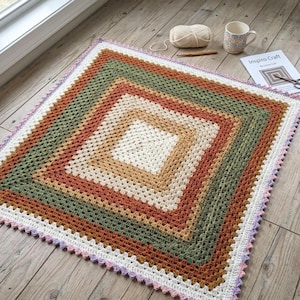 May include: A square crochet blanket featuring concentric layers of cream, tan, rust orange, and olive green yarn. The blanket has a scalloped border in light purple and white. Nearby are a ball of cream yarn, a crochet hook, and a mug.