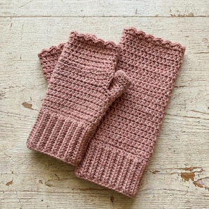 Op de afbeelding: Een paar stoffig roze vingerloze handschoenen en een bijpassende polswarmers, beide met de hand gehaakt. De handschoenen hebben een geribbelde manchet en een geschulpte rand. De polswarmers zijn rechthoekig met een geschulpte rand. De items worden gepresenteerd op een licht houten oppervlak.