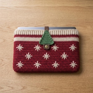May include: A rectangular crochet laptop sleeve in deep red featuring a white snowflake pattern. The top edge has a cream-colored ribbed border and a central brown tab with a green crochet Christmas tree shape and a gold-toned metal snap button.