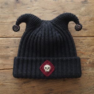 Pode incluir: Um gorro de crochê preto com duas protuberâncias pontiagudas em forma de chifre no topo, cada uma terminando em um pequeno detalhe em espiral. A frente apresenta um emblema em forma de losango vermelho escuro com um desenho de caveira branca.