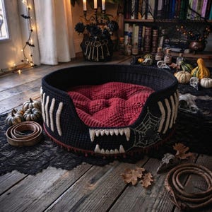May include: A round, black crocheted pet bed featuring a deep red tufted interior cushion. The exterior is decorated with white crocheted rib-like accents, a set of sharp white fangs at the front, and a detailed spiderweb pattern on the side.