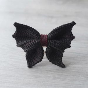 May include: A black crocheted bow with bat-wing shaped edges and a dark maroon center wrap. The accessory features a textured yarn construction with pointed wing tips on both sides, creating a symmetrical silhouette that resembles a bat in flight.