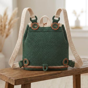 May include: A dark green crochet backpack featuring cream-colored shoulder straps and a top handle. The bag is accented with circular rust-colored rings at the strap attachment points and a matching rust-colored horizontal stripe near the base.