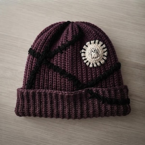 May include: A dark purple ribbed knit beanie featuring thick, black yarn accents stitched in a crisscross pattern across the surface. A circular, cream-colored crocheted patch is attached to the side, displaying a small, embroidered ghost figure.