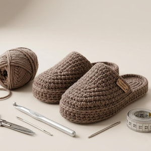 Peut inclure: Une paire de pantoufles crochetées à la main, de couleur marron. L'image comprend une pelote de laine, un crochet, des ciseaux, des aiguilles et un mètre ruban. Les pantoufles ont une petite étiquette qui dit "Handmade Crochet Slippers".