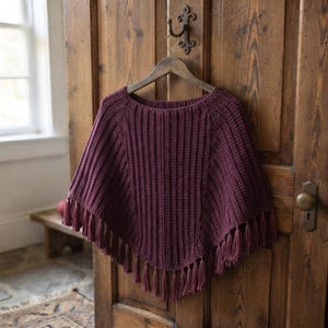 May include: A dark plum-colored crochet poncho featuring a vertical ribbed texture throughout the body. The garment has a wide neckline and a pointed hemline finished with long, matching yarn tassels hanging from the bottom edge.